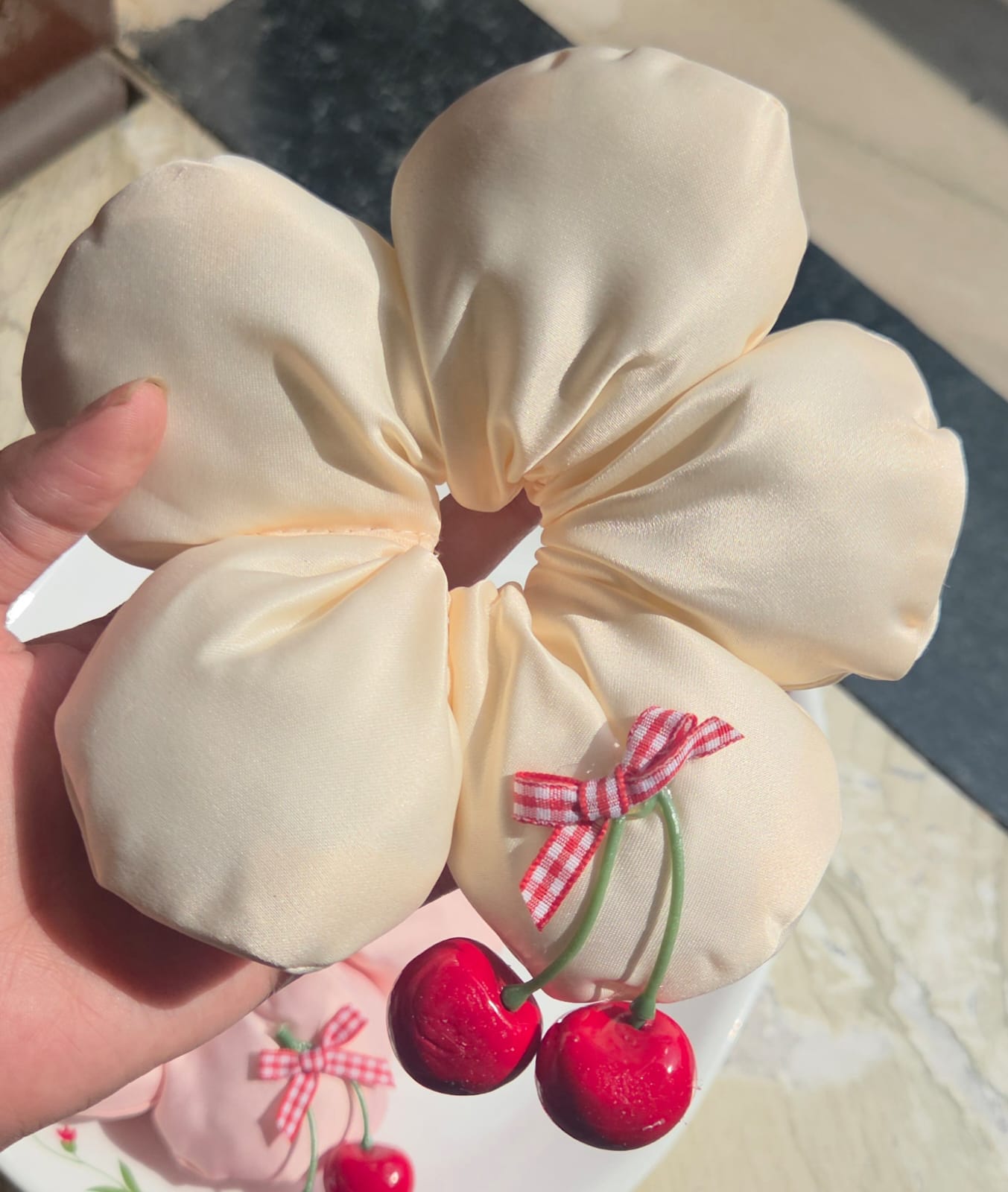 A soft cream-colored ivory satin flower bun scrunchie with a mini red gingham ribbon and hanging cherry detail.