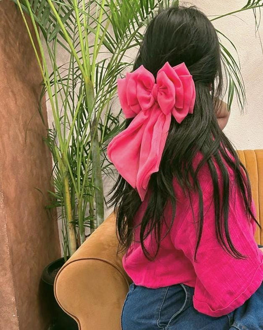 Rear view of a woman with long, dark wavy hair wearing a large, oversized bright pink georgette hair bow clip. The bow features elegant, long cascading fabric tails that drape down her hair. She is dressed in a matching pink top and blue jeans, sitting indoors next to a green potted palm plant.