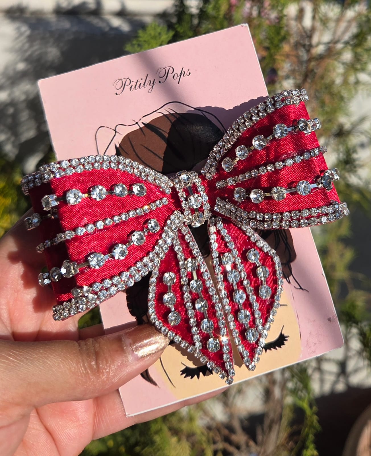 Close-up of a vibrant ruby red satin bow hair clip accented with rows of clear crystal rhinestones on a pink Pitily Pops card.