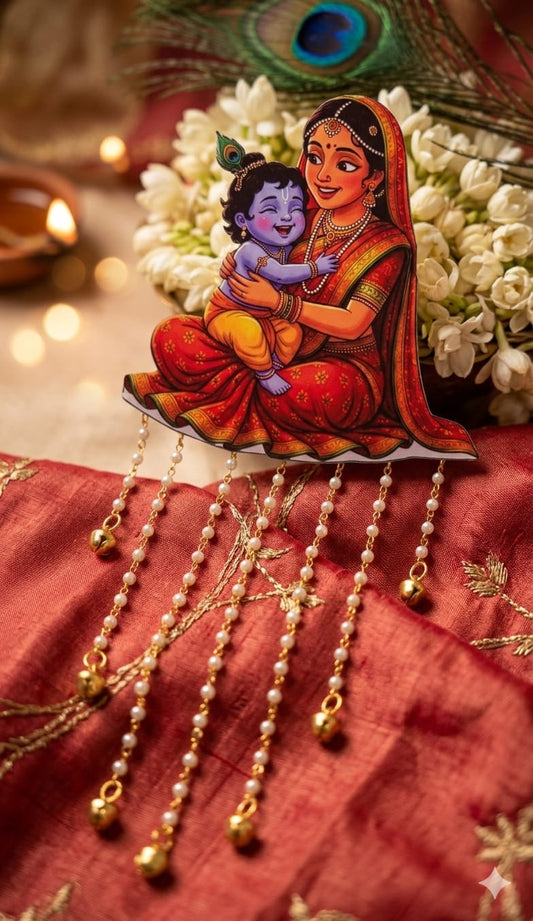 ​A close-up shot of a traditional Indian hair accessory featuring a colorful illustration of Yashoda holding baby Krishna. The accessory is placed on a textured red and gold silk fabric, with white jasmine flowers (mogra), a peacock feather, and a lit diya blurring softly in the background. Pearl-beaded strands with small golden ghungroo bells hang elegantly from the base