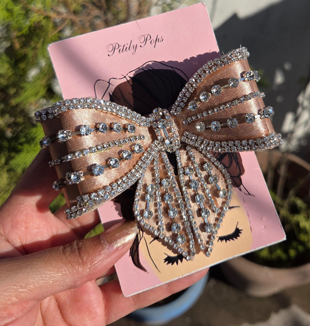 Handcrafted champagne gold satin hair bow covered in rows of sparkling crystal rhinestones on a Pitily Pops brand card.