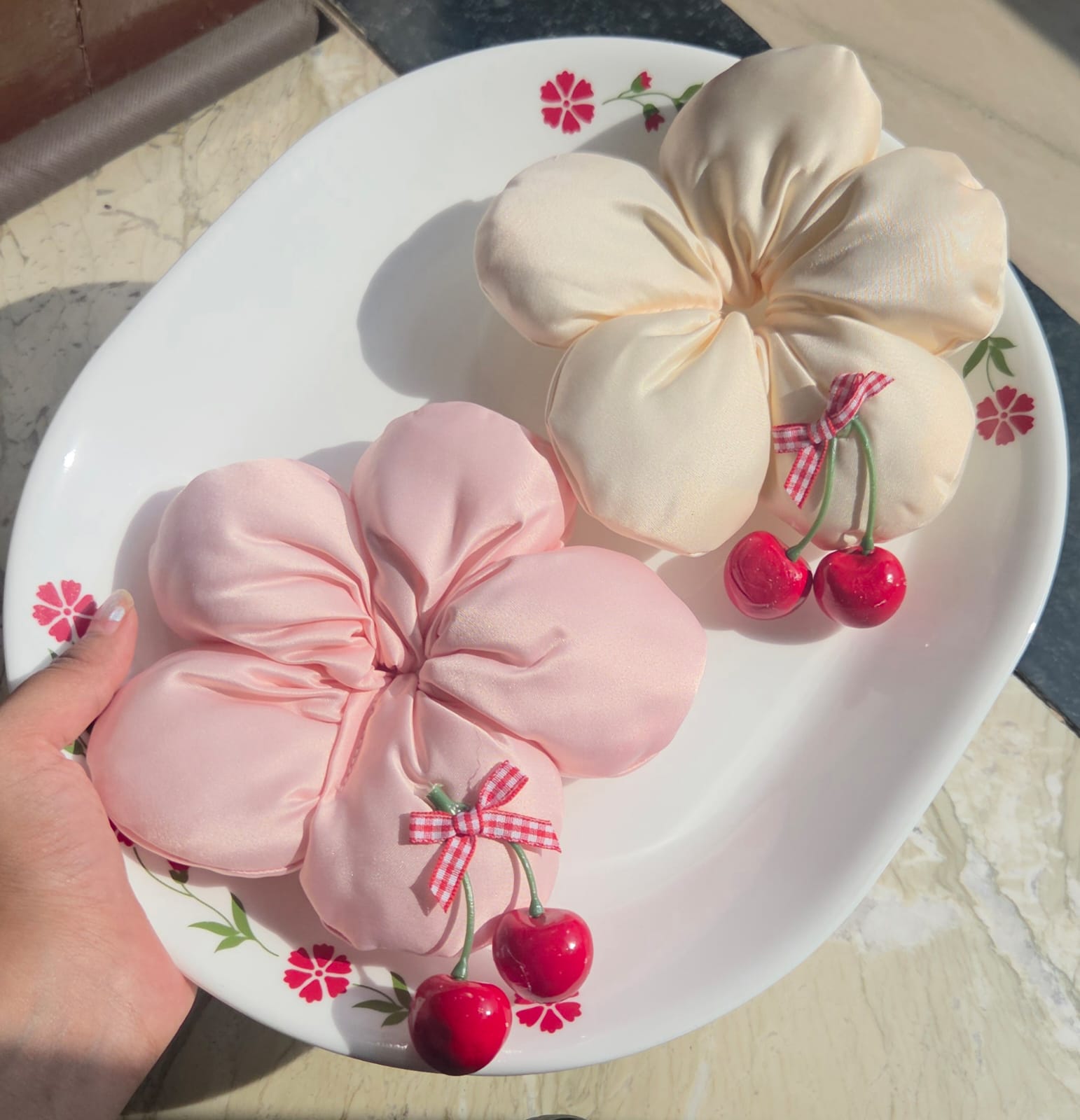 Two oversized handmade flower bun scrunchies in blush pink and cream satin fabric, featuring dangling red cherry charms, displayed on a decorative plate.
