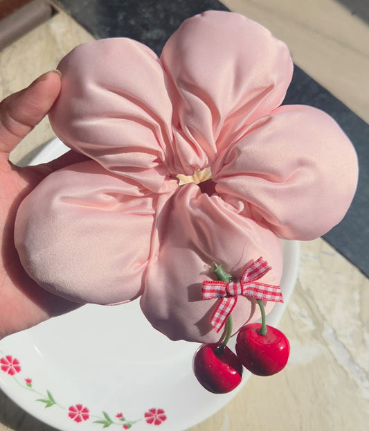 Close-up of a handmade blush pink satin flower scrunchie with five large petals and a red cherry accent charm.