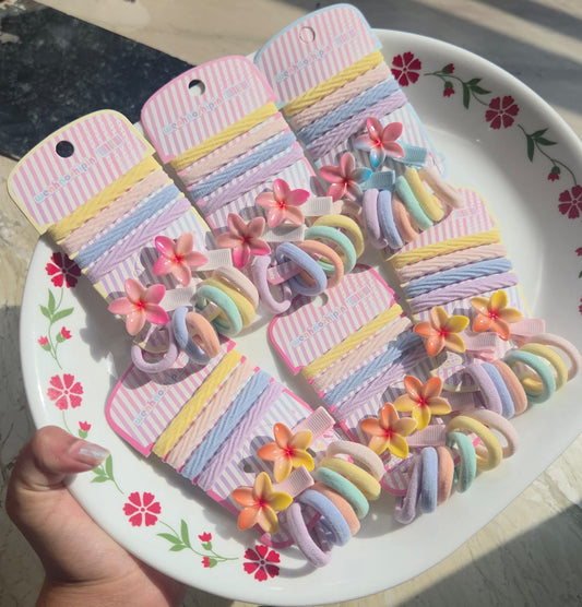 A collection of PitilyPops pastel floral hair clip and elastic band sets arranged on a white decorative floral plate, showing pink and yellow color options.