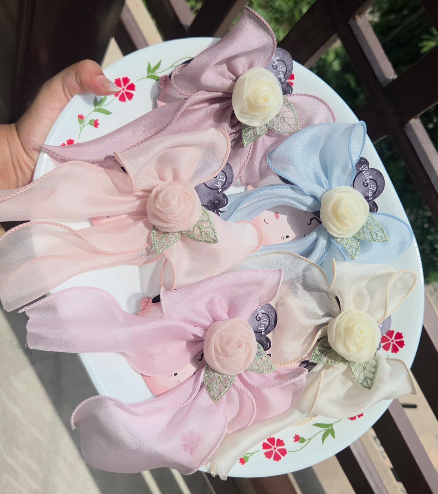 Five variations of sheer organza hair bows with central rose accents and alligator clips in pastel shades of pink, mauve, blue, cream, and peach