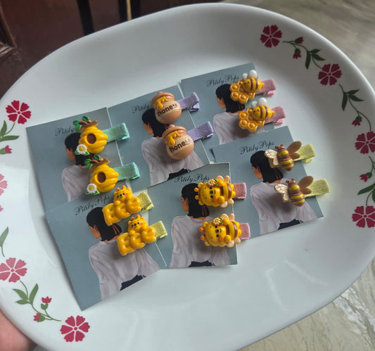 Six pairs of handmade kids bee and honey themed hair clips by Pitily Pops displayed on a white floral plate