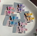 Five pairs of Pitily Pops handmade kids butterfly hair clips in shimmering pastel shades of pink, purple, blue, yellow, and iridescent white, displayed on branded cards showing fabric-lined alligator clips