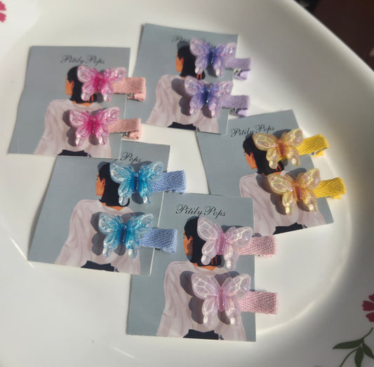 Five pairs of Pitily Pops handmade kids butterfly hair clips in shimmering pastel shades of pink, purple, blue, yellow, and iridescent white, displayed on branded cards showing fabric-lined alligator clips