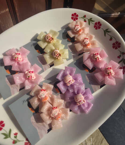 Four pairs of kids hair clips in pink, purple, yellow, and peach colors, featuring sparkly sheer ribbon bows with cute cartoon charms, displayed on backing cards inside a white dish 