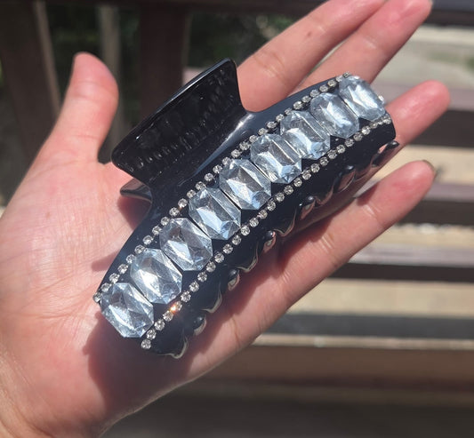    A close-up view of a woman's hand holding a high-shine black premium hair clutcher adorned with emerald-cut crystals and tiny sparkling rhinestones.
