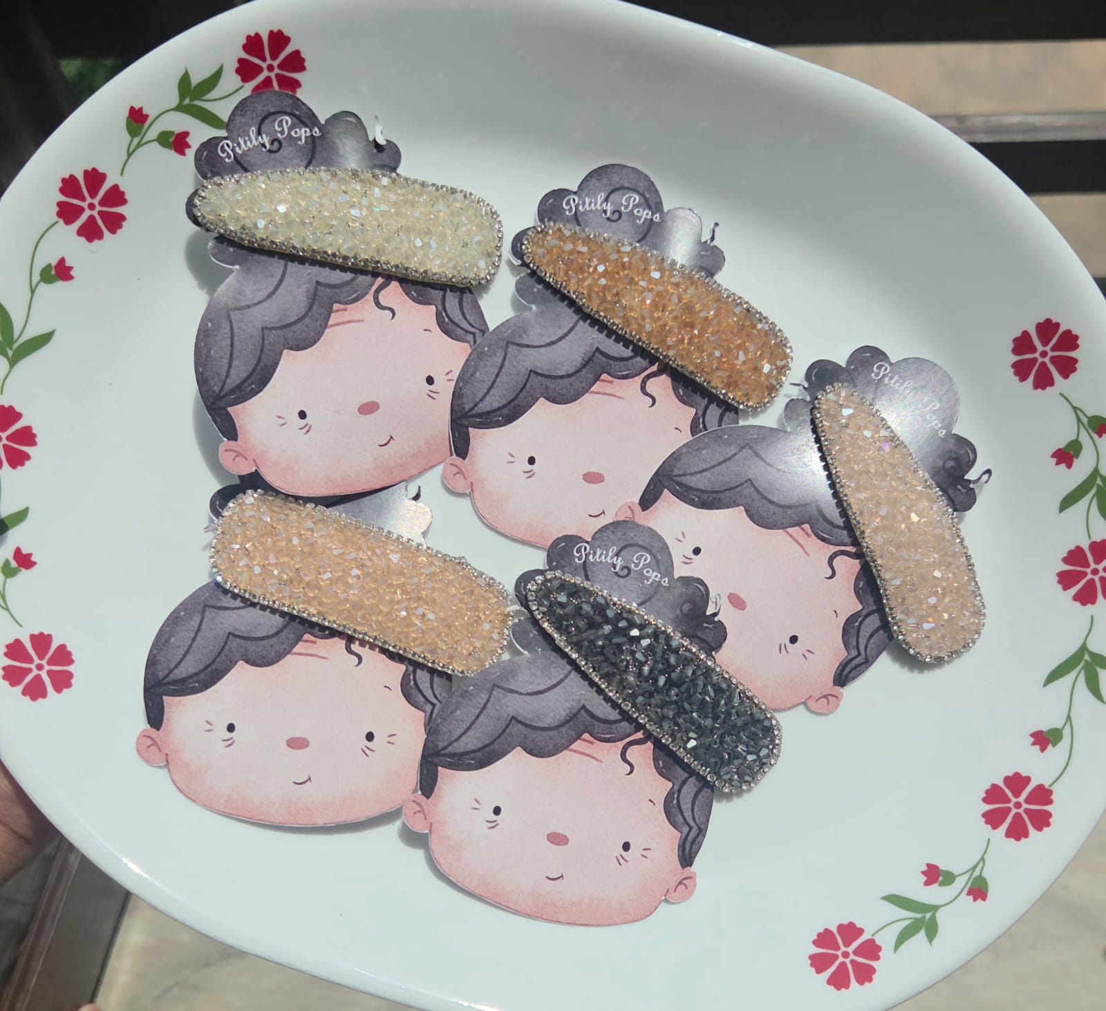 Five sparkling Pitily Pops crystal rhinestone hair clips in white, gold, blush pink, beige, and charcoal black arranged on custom cartoon backing cards.
