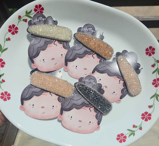 Five sparkling Pitily Pops crystal rhinestone hair clips in white, gold, blush pink, beige, and charcoal black arranged on custom cartoon backing cards.
