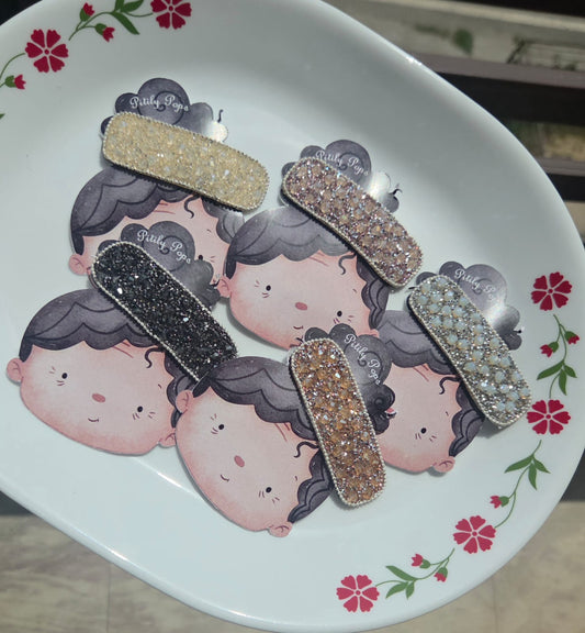 Five sparkly rhinestone crystal rectangular hair barrettes in champagne gold, rose gold, black, opal white, and amber bronze displayed on a decorative plate.
