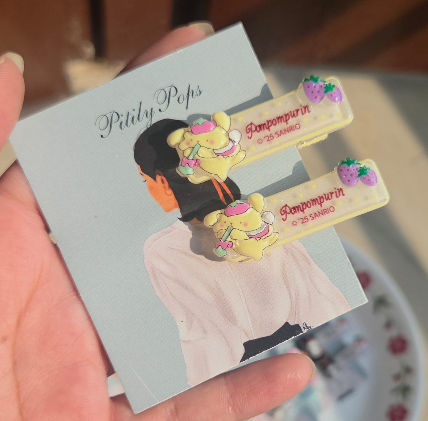 Pair of pastel yellow kids alligator hair clips featuring the cute cartoon character Pompompurin with small purple berry charms.