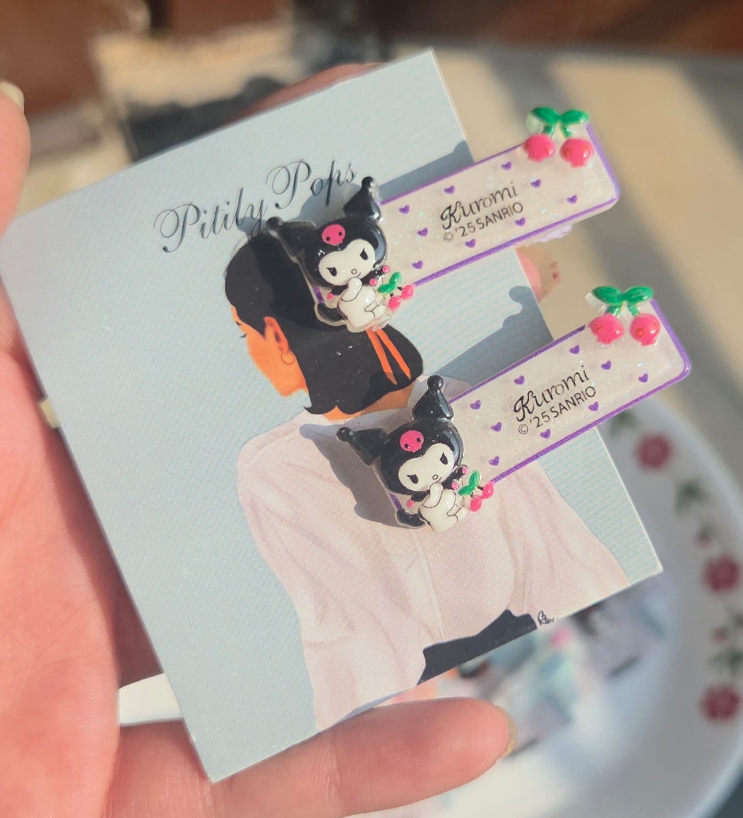 Pair of lavender purple pastel hair clips for kids featuring the cheeky cartoon character Kuromi with red cherry details.