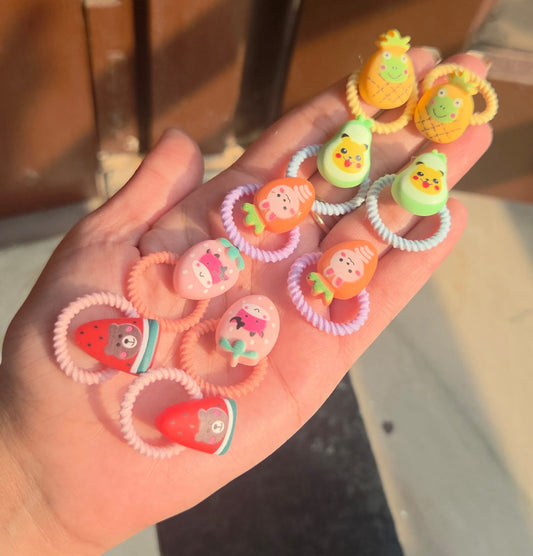 Close up of five pairs of matching toddler elastic hair bands featuring cute pineapple, avocado, strawberry, carrot, and watermelon designs.