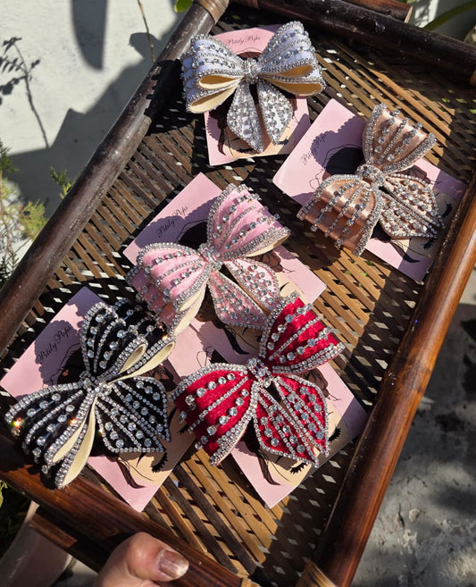 A woven bamboo tray held outdoors containing five sparkly, rhinestone-encrusted bow hair clips from Pitily Pops in white, beige, light pink, black, and deep red