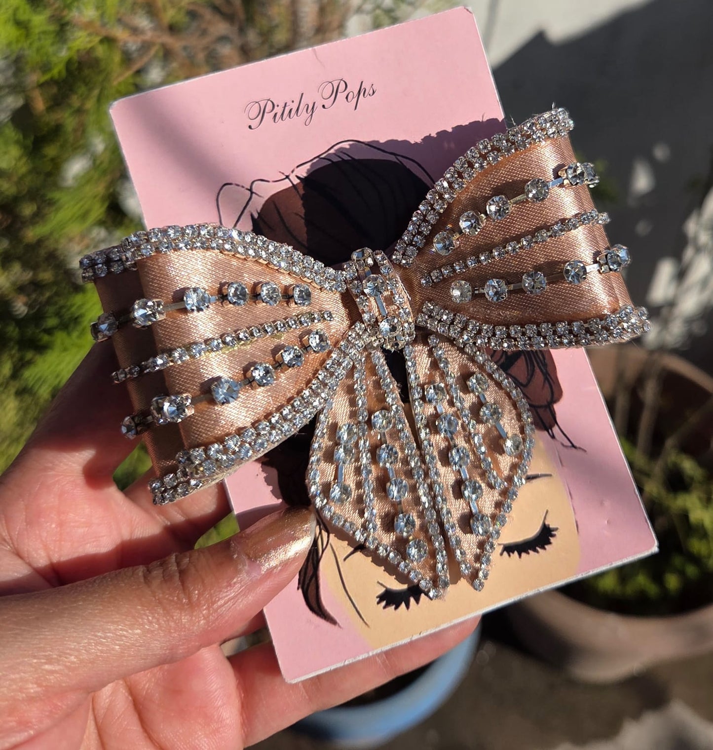 Close-up of a handmade beige satin bow hair clip heavily embellished with rows of glittering rhinestones on a pink Pitily Pops card.