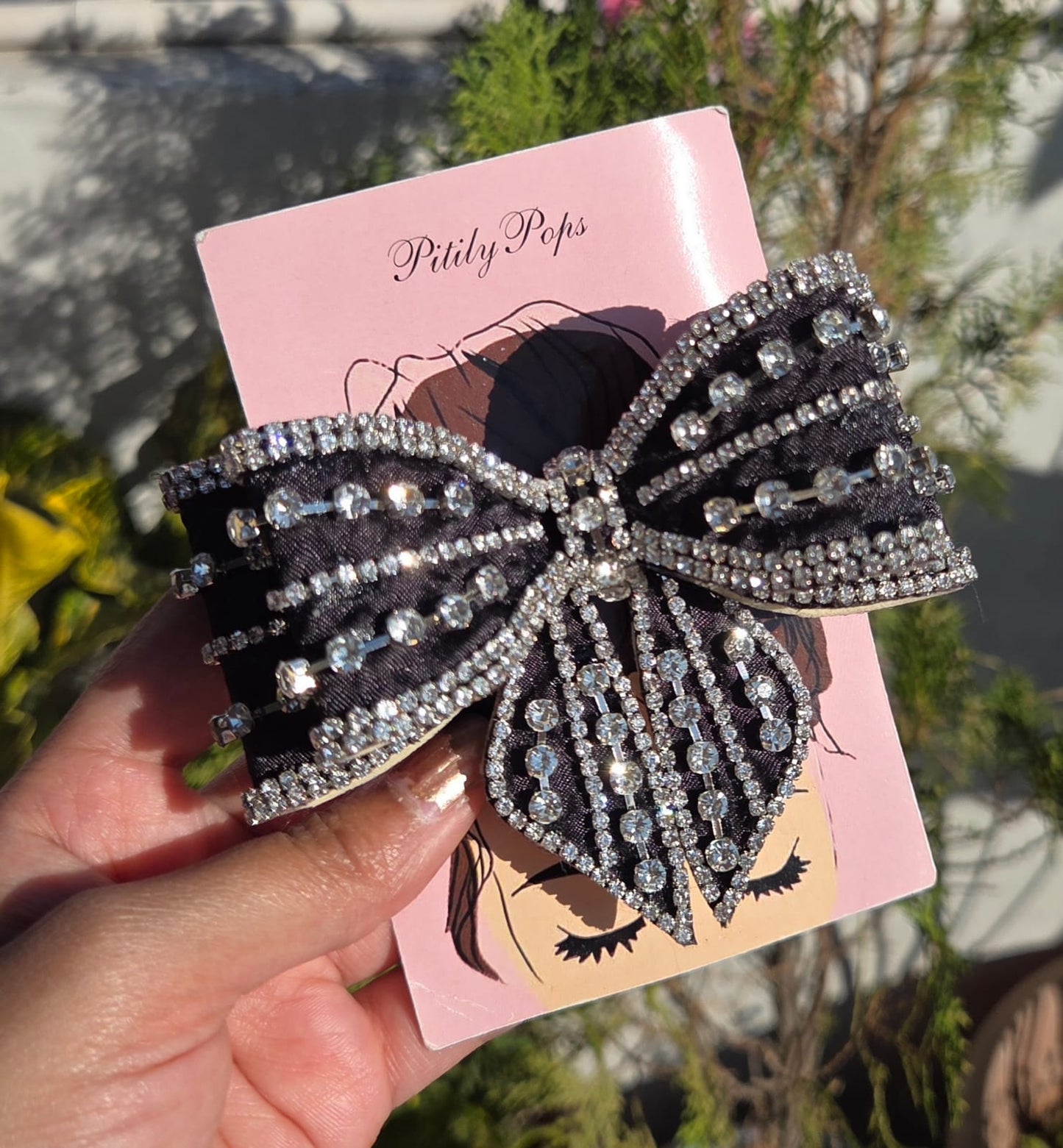 Close-up of a black satin bow hair clip embellished with shimmering rows of clear rhinestones on a branded Pitily Pops card.