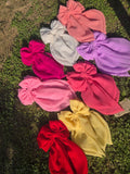 An overhead flat lay shot of eight oversized handmade georgette fabric hair bows with long tails, arranged together on a patch of green grass under natural sunlight. The color collection features white, lavender, peach, magenta, coral pink, bright pink, pastel yellow, and classic red.