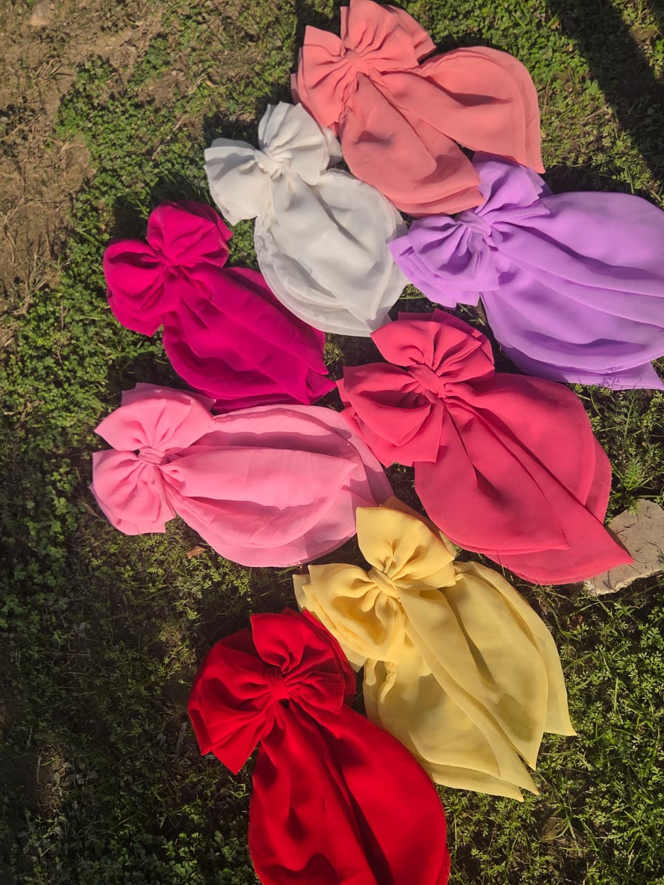 An overhead flat lay shot of eight oversized handmade georgette fabric hair bows with long tails, arranged together on a patch of green grass under natural sunlight. The color collection features white, lavender, peach, magenta, coral pink, bright pink, pastel yellow, and classic red.
