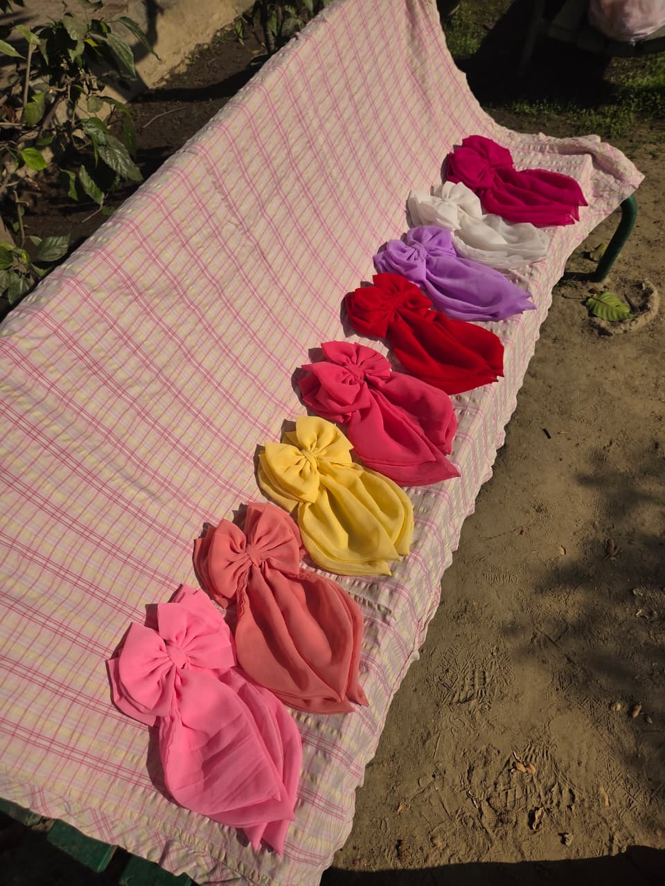 ​A lineup of eight oversized georgette hair bow clips with long draped tails, displayed in a row on a pink and white plaid blanket covering an outdoor bench. The vibrant collection showcases a gradient of colors from light pink, peach, yellow, and red, to magenta, lilac, white, and deep pink under bright daylight.