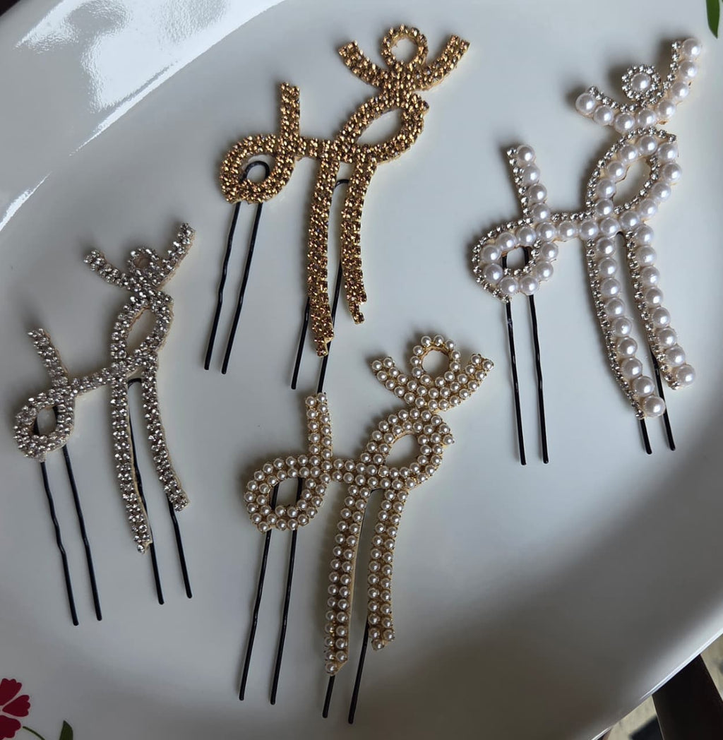 Flat lay display of four different variants of a traditional Devanagari "Maa" script hair pin on a white ceramic plate. The collection includes a silver rhinestone version, a gold rhinestone version, a full seed pearl version, and a mixed pearl and crystal bordered design, each attached to black u-shaped hair pins.