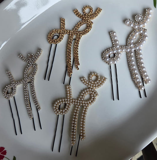 Flat lay display of four different variants of a traditional Devanagari "Maa" script hair pin on a white ceramic plate. The collection includes a silver rhinestone version, a gold rhinestone version, a full seed pearl version, and a mixed pearl and crystal bordered design, each attached to black u-shaped hair pins.