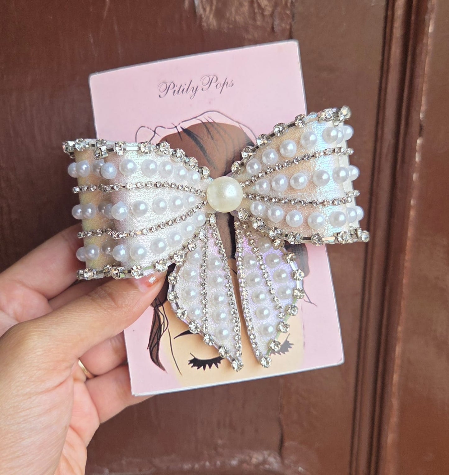 Oversized white pearl and crystal rhinestone bow hair clip on branded Pititly Pops pink card held in hand outdoors