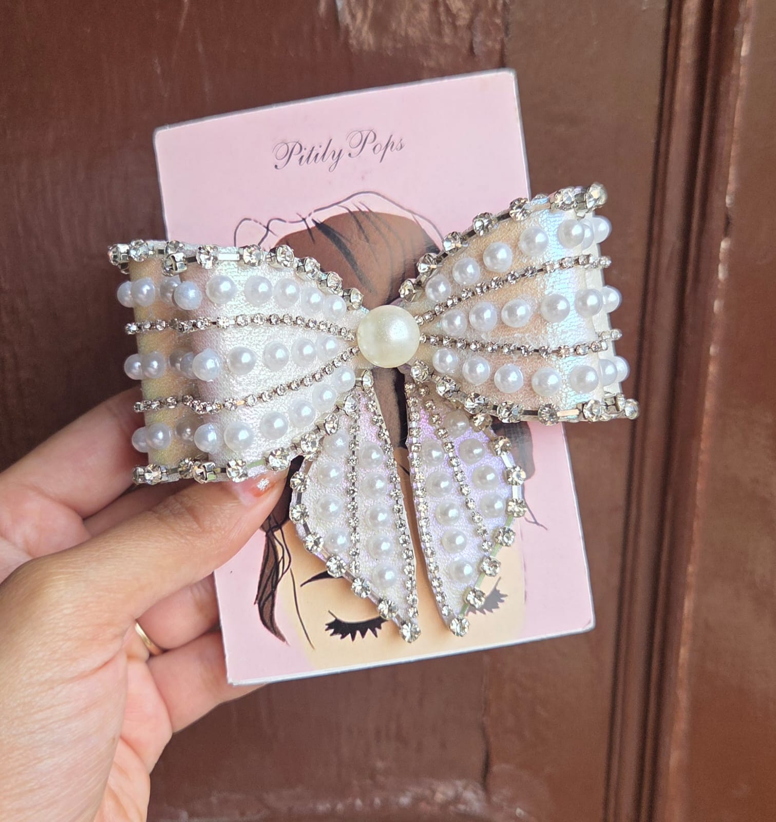 Oversized white pearl and crystal rhinestone bow hair clip on branded Pititly Pops pink card held in hand outdoors