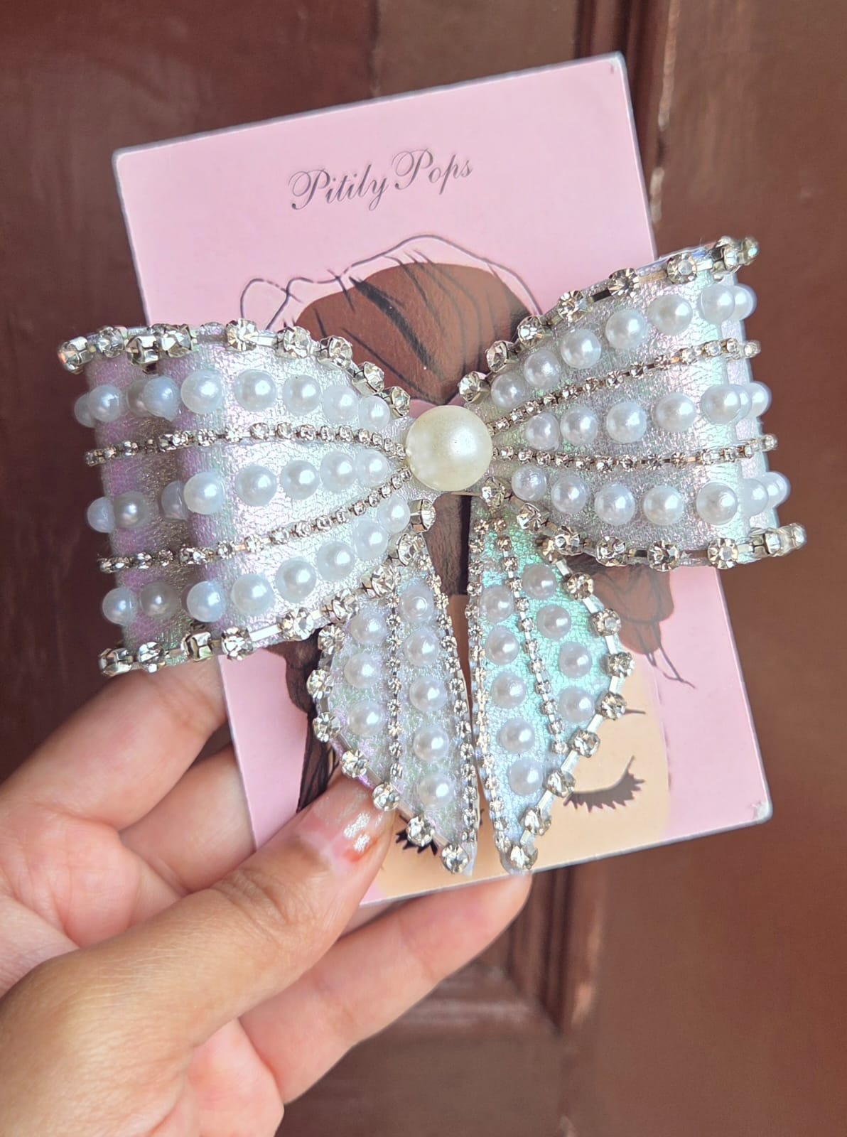 Pitily Pops shimmery sky blue pearl and rhinestone bow barrette hair clip held up for display, showing layered bow design with buckle back
