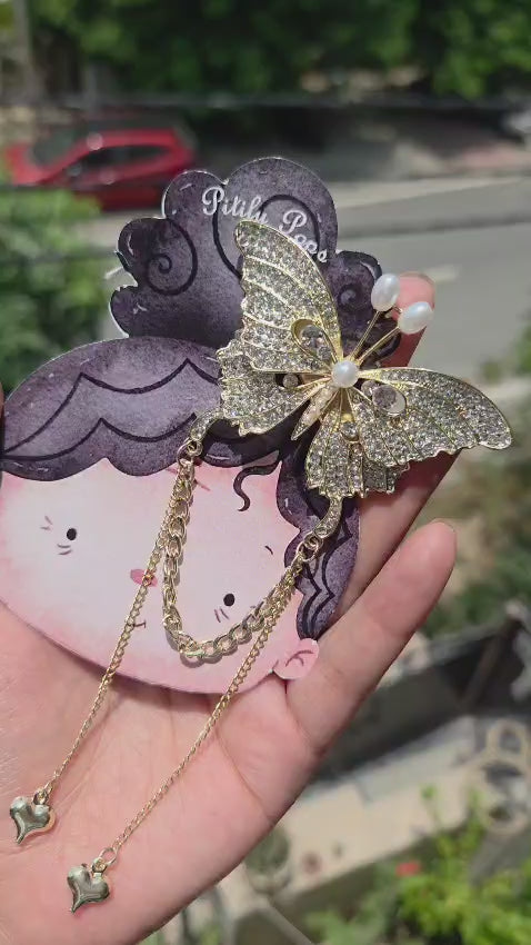 A hand holding a gold anti-tarnish butterfly hair clip with shimmering rhinestones, pearl details, and dangling gold chains with heart charms.