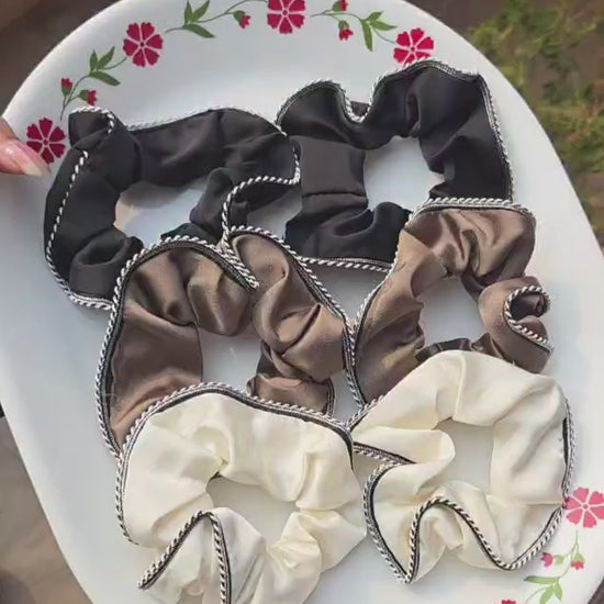 A collection of six premium ruffled satin scrunchies with black and white contrast piped edges, displayed on a decorative white plate. The collection features three color options: two classic black, two coffee brown, and two creamy off-white hair ties.