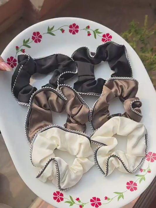 A collection of six premium ruffled satin scrunchies with black and white contrast piped edges, displayed on a decorative white plate. The collection features three color options: two classic black, two coffee brown, and two creamy off-white hair ties.