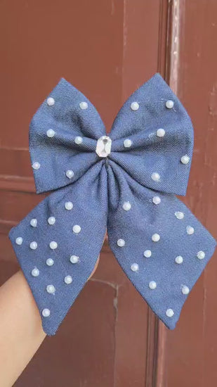 Oversized denim hair bow accessory with pearl and stone work details, held against a wooden background.