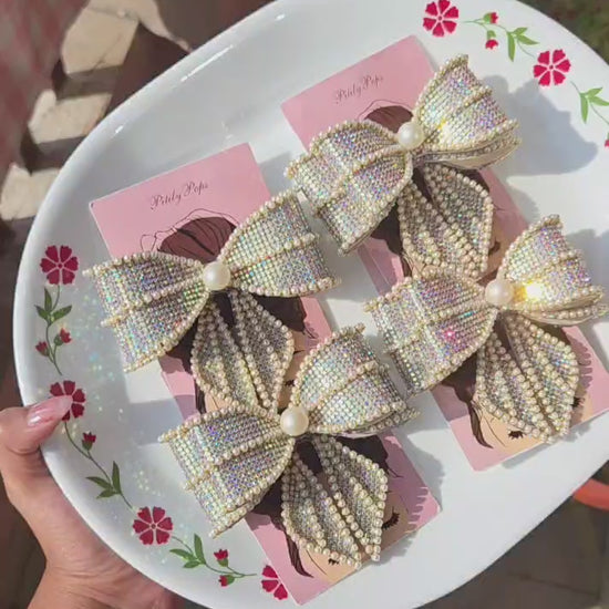 Rhinestone and pearl embellished hair bow clips with buckle barrette back, displayed on a floral white plate — bridal and party hair accessory