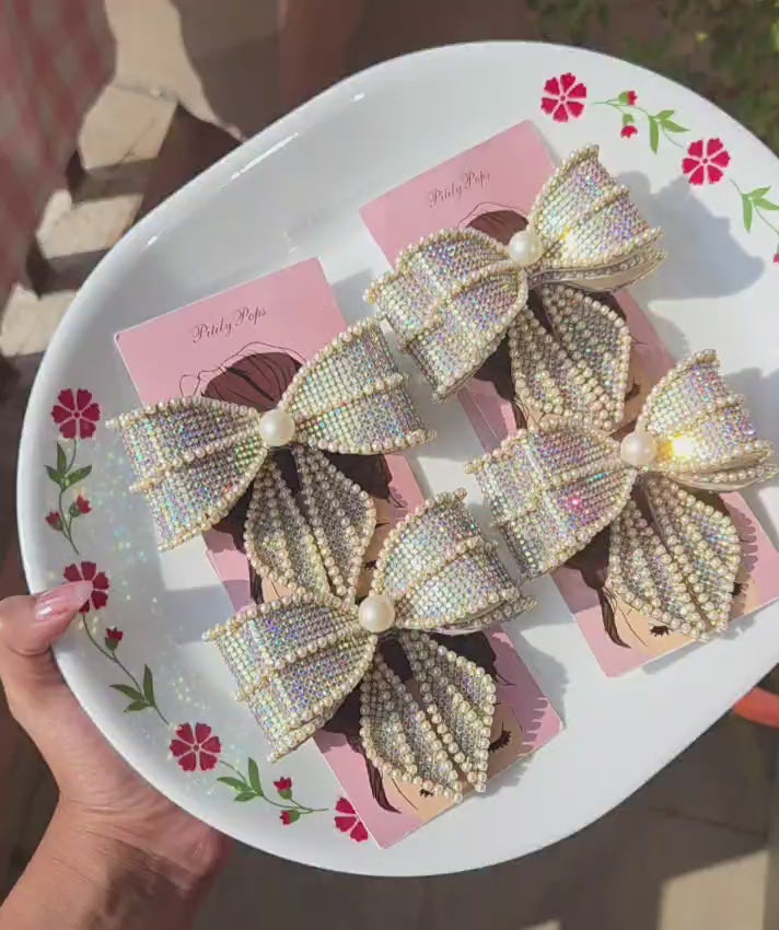 Rhinestone and pearl embellished hair bow clips with buckle barrette back, displayed on a floral white plate — bridal and party hair accessory