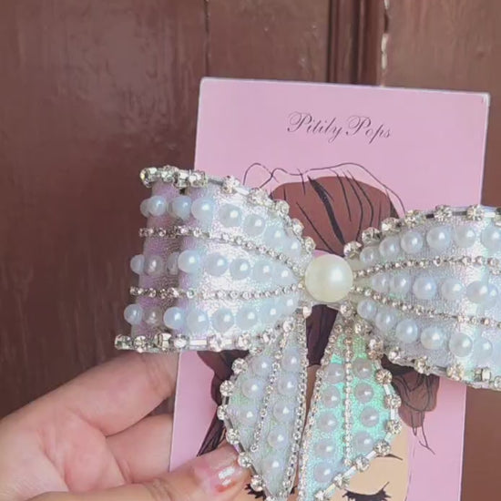 Close-up of a glamorous oversized bow hair clip covered in white faux pearls and sparkling rhinestone chain trim on iridescent sky blue shimmer fabric, secured with a barrett