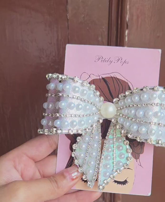 Close-up of a glamorous oversized bow hair clip covered in white faux pearls and sparkling rhinestone chain trim on iridescent sky blue shimmer fabric, secured with a barrett