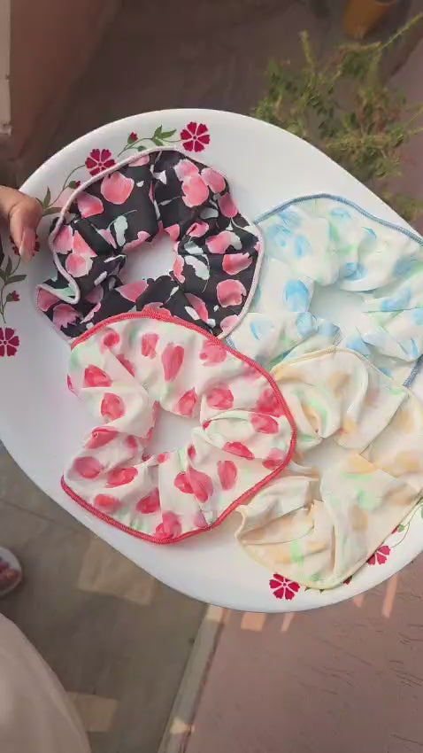 Pitily Pops handmade floral fabric scrunchies in four pastel color variants displayed on a white plate.