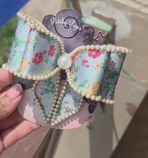 Blue floral satin hair bow with pearl bead trim and alligator clip, held on a decorative display card