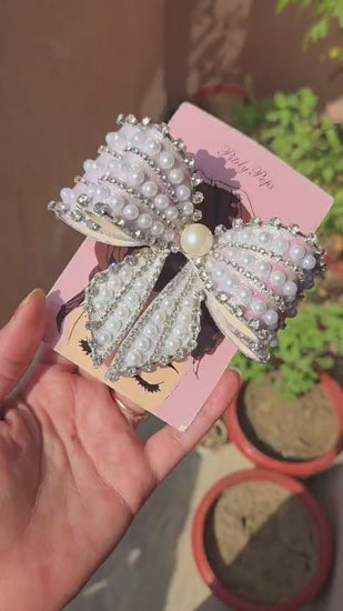 Close-up of large decorative hair bow with faux pearls, rhinestone trim, and flowing tails on pink brand card — elegant women's hair accessory